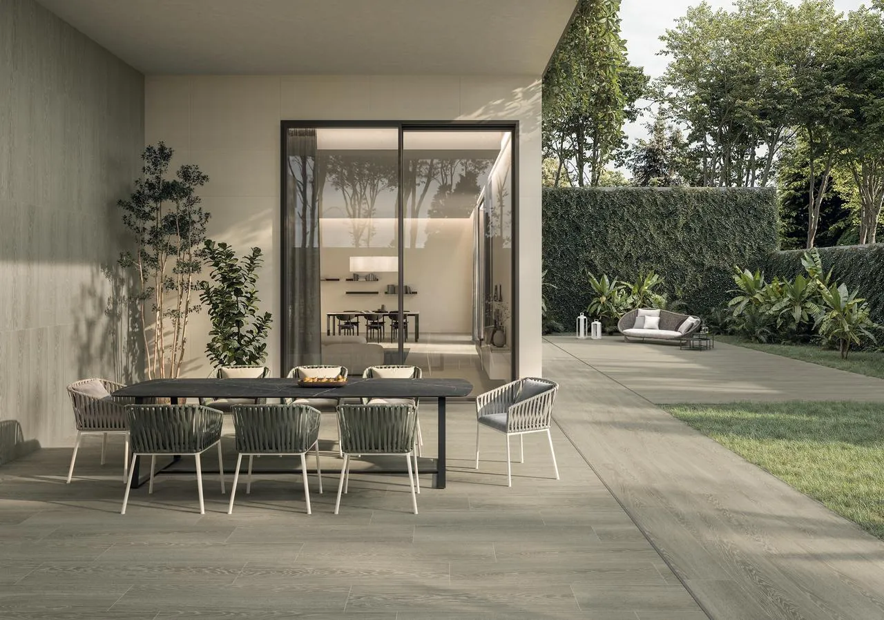 Giardino con una grande finestra che da su un patio in gres Atlas Concorde, un tavolino e diverse sedie, nel verde