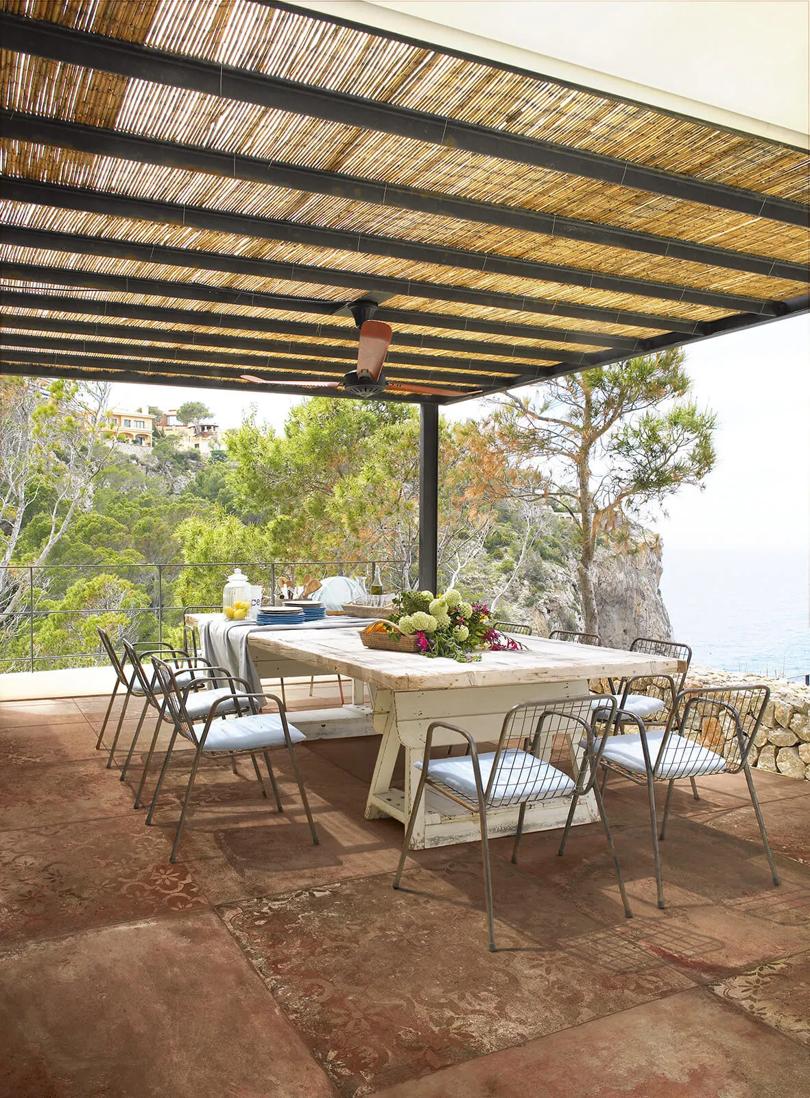 Patio di una casa con pavimento in cotto tagina, un grande tavolino e sedie sotto una tettoia in rafia, in mezzo al verde