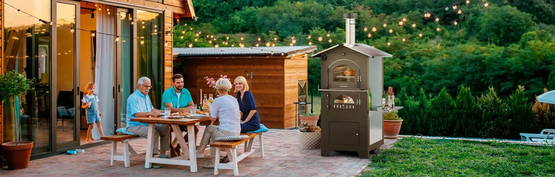 Esterno di una casa in legno, in giardino una famiglia di 2 anziani, 2 giovani e una bimba, e seduta ad un tavolo con a fianco un forno Gusto a legna Fontana acceso, in mezzo al verde