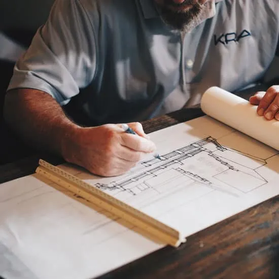 Uomo con barba brizzolata e camicia disegna un progetto ad un tavolo di legno